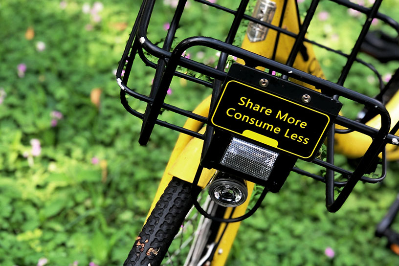 Ein gelbes Fahrrad im Grünen. Am Korb steht ein Schild mit der Aufschrift: "Share more, Consume less". (Mehr Teilen, Weniger Verbrauchen)