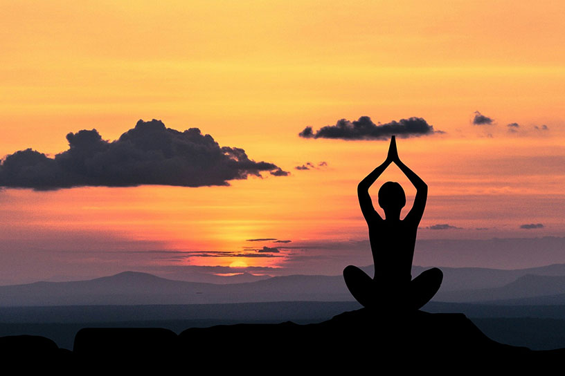 Silouette eines Menschen der Meditiert. Im Hintergrund sieht man einen orang/roten Sonnenuntergang zwischen fernen Bergen.