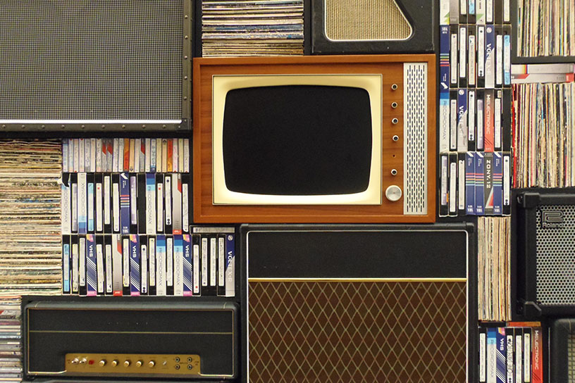 Alter Fehrnseher und Videokasetten in einem Regal, umgeben von alten Lautsprechern.