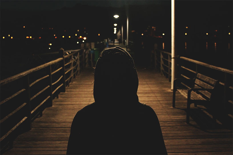 Mensch mit dem Rücken zum Betrachter, auf einem Holzsteg bei Nacht. Alles ist dunkel bis auf die Straßenlaternen, die den Steg anleuchten.