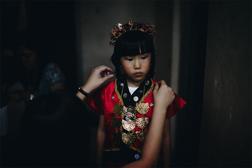 Ein asiatisches Mädchen wird ein traditionelles rotes Kleid angezeogen von Händen aus dem Off. Das Mädchen schaut etwas traurig nach rechts weg.