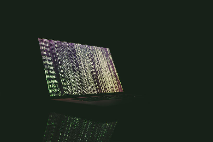 Ein Laptop in einem komplett schwarzen Raum. Über den Bildschirm laufen grün, gelbe Zeichenreihen von oben nach unten.
