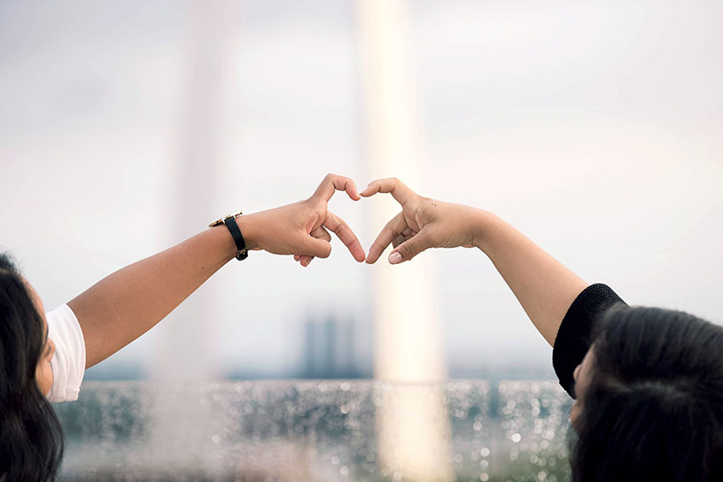 Zwei Menschen formen ein Herz mit ihren Händen. Der Hintergrund ist verschwommen und man erkennt einen Regenbogen sowie Glasscheiben mit Regentropfen.